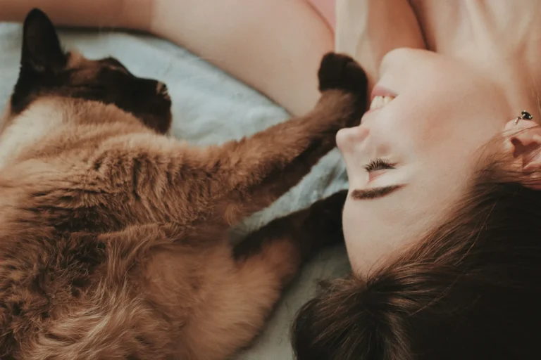A Siamese cat lies next to a woman, with its dark paw gently touching her face as they look toward each other.